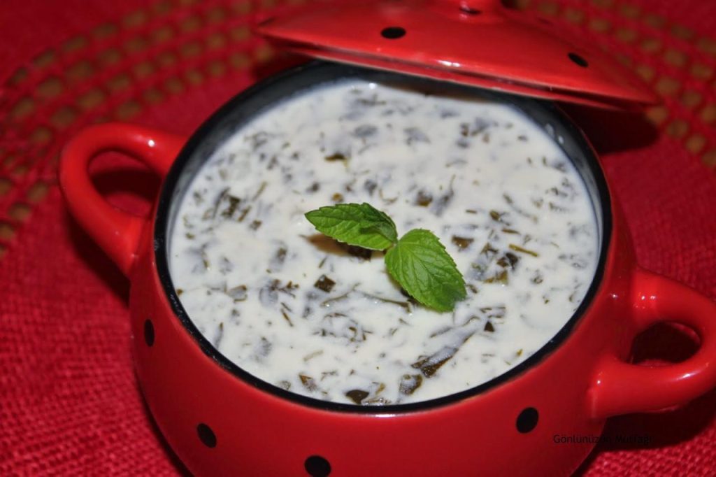 Chilled yogurt soup presented in a vibrant red, polka-dotted bowl placed on a textured red tablecloth. A fresh mint sprig adds a touch of green.