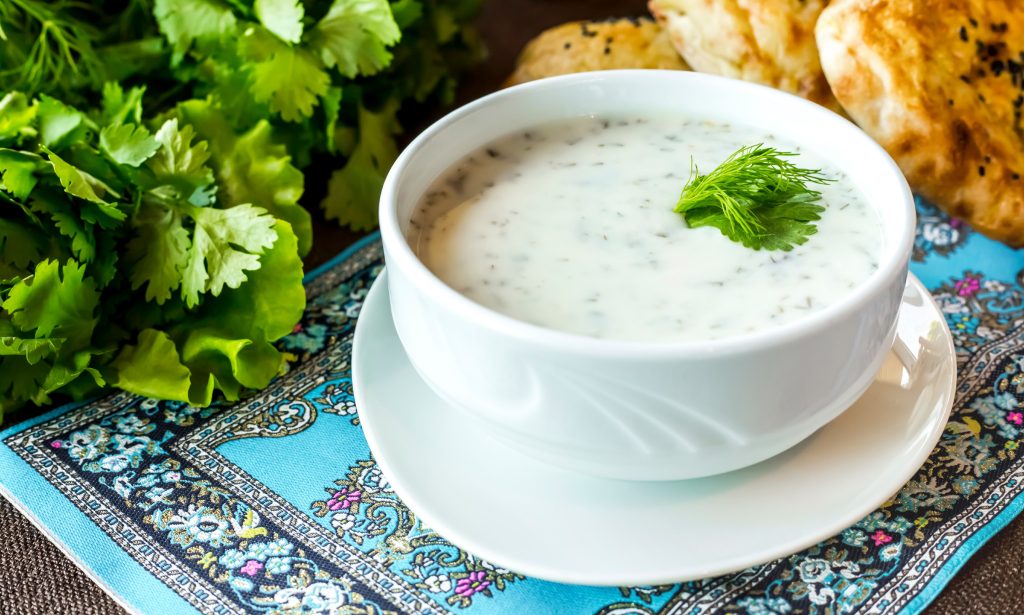 Cold yogurt and dill soup served in a sleek white bowl on a decorative blue placemat. Fresh herbs and flatbread are visible in the background.