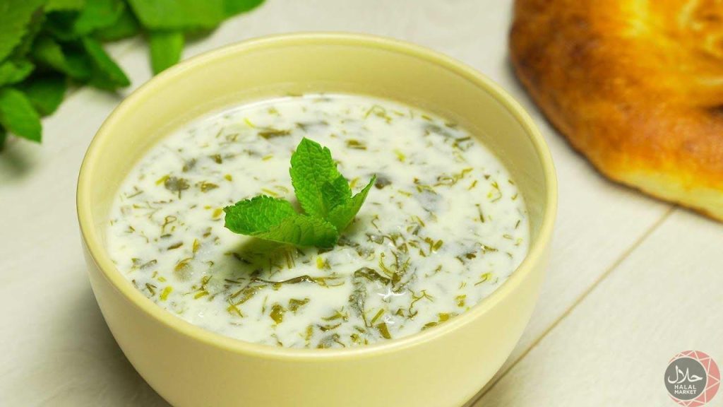 Creamy Azerbaijani Dovga, a yogurt-based soup, served in a pale yellow bowl on a light wooden table. Fresh mint sprigs garnish the soup.