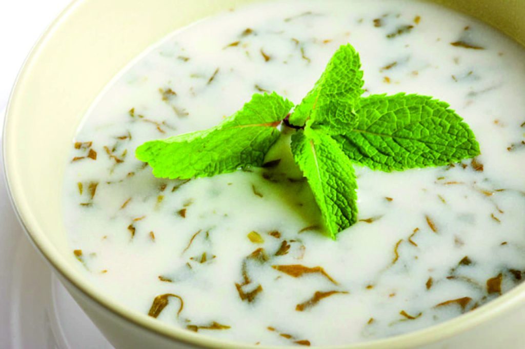 Close-up of a creamy yogurt and herb soup in a light-colored bowl. The soup is topped with fresh mint leaves, highlighting its vibrant green color.