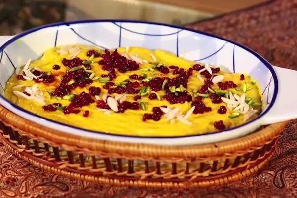 A bowl of Persian yogurt stew with a golden-yellow hue, topped with slivers of pistachios and barberries for decoration.