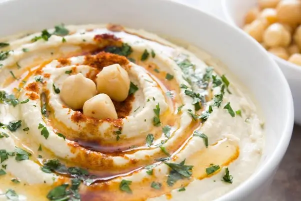 Smooth-hummus-dip-garnished-with-whole-chickpeas-fresh-herbs-olive-oil-and-paprika-in-white-bowl