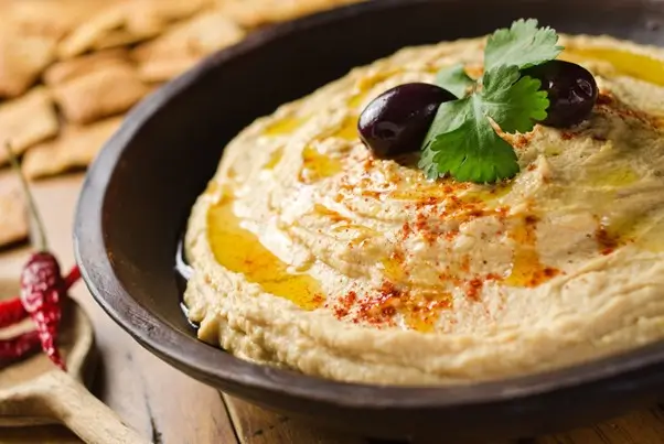 Traditional-hummus-dip-topped-with-black-olives-cilantro-olive-oil-and-paprika-in-dark-bowl