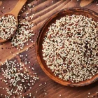 A bowl of cooked quinoa with a garnish and a bowl of raw quinoa grains, placed on a wooden surface with a spoon