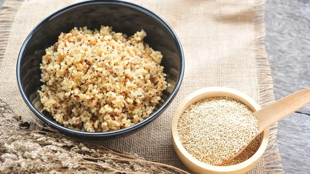 A bowl of fluffy cooked quinoa with a small wooden bowl of raw quinoa grains placed beside it.