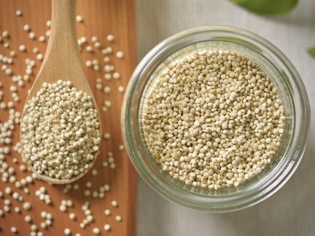 A glass jar filled with white quinoa grains, with a wooden spoon containing quinoa placed on a wooden board.